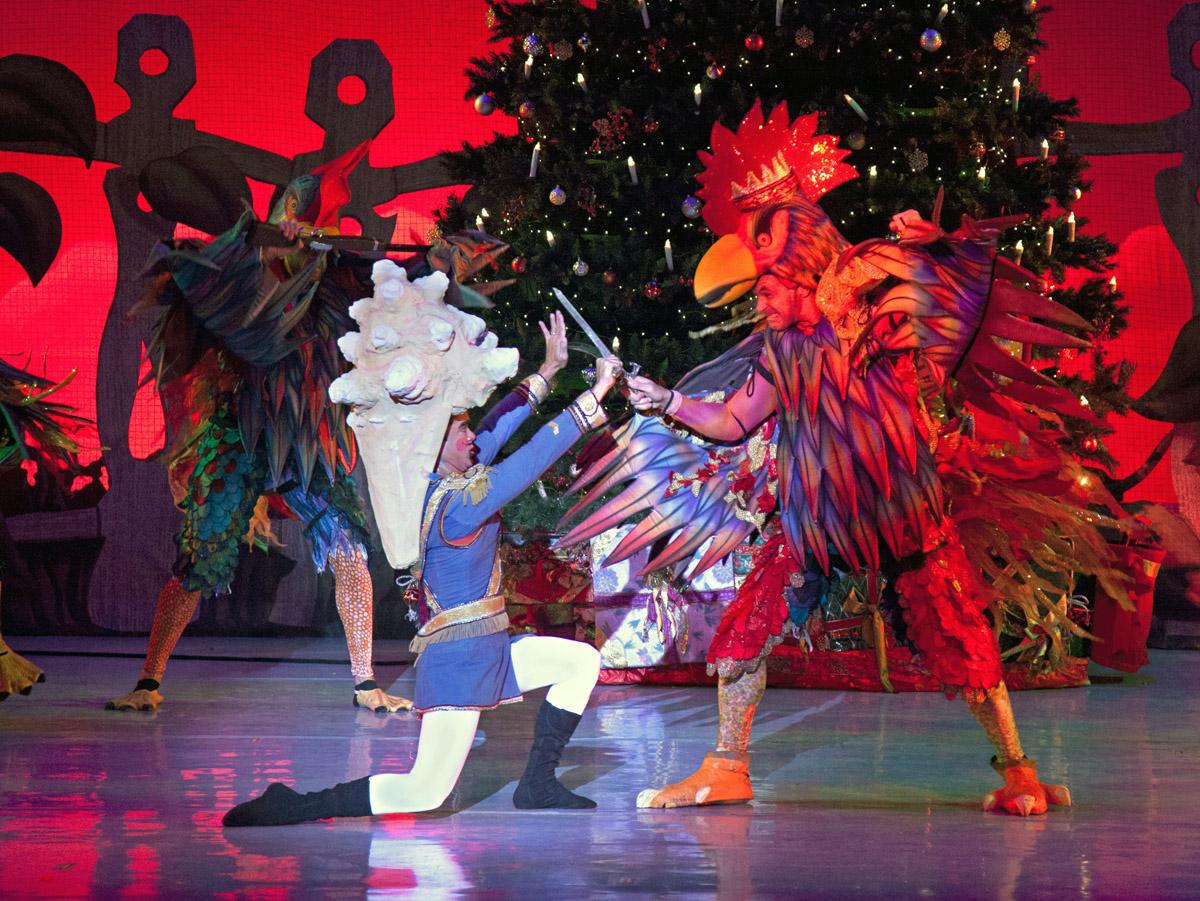 Dancers in colorful feathered costumes perform on a brightly lit stage.