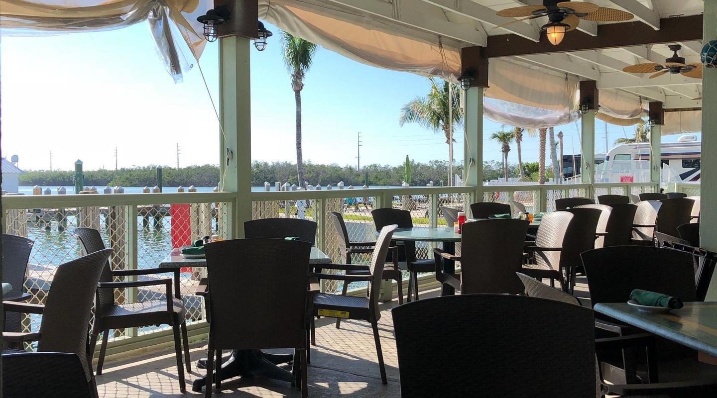 Outdoor waterfront restaurant patio with empty black chairs and tables under a canopy.