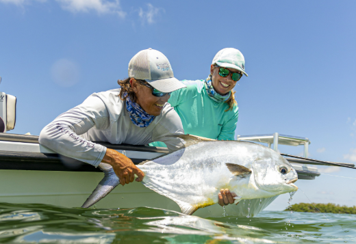 Two anglers release a large fish beside a boat on calm water.