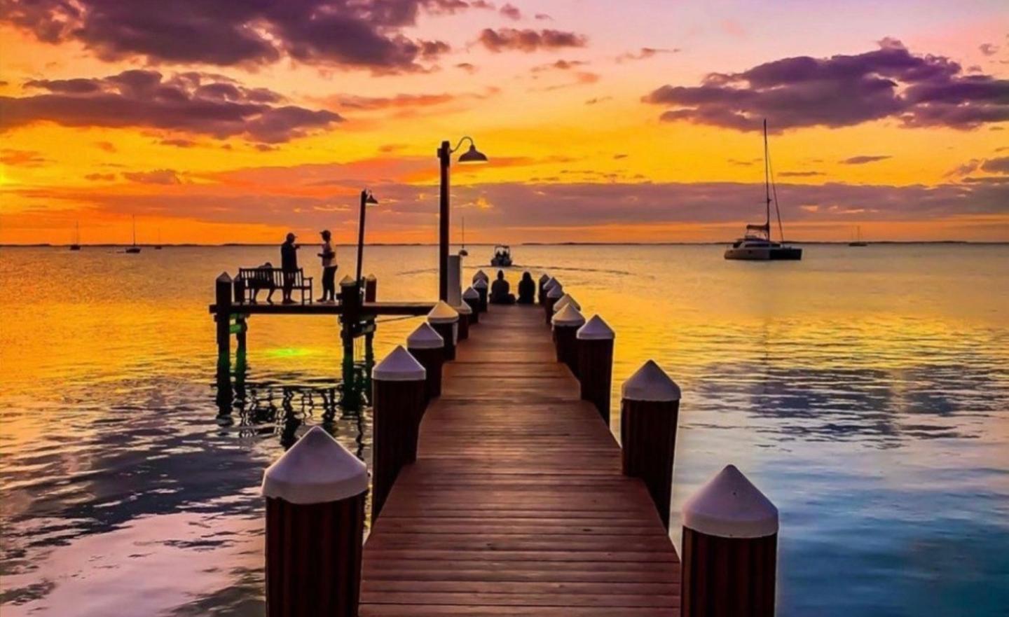 Wooden pier leading into calm water at sunset, with sailboats and glowing clouds
