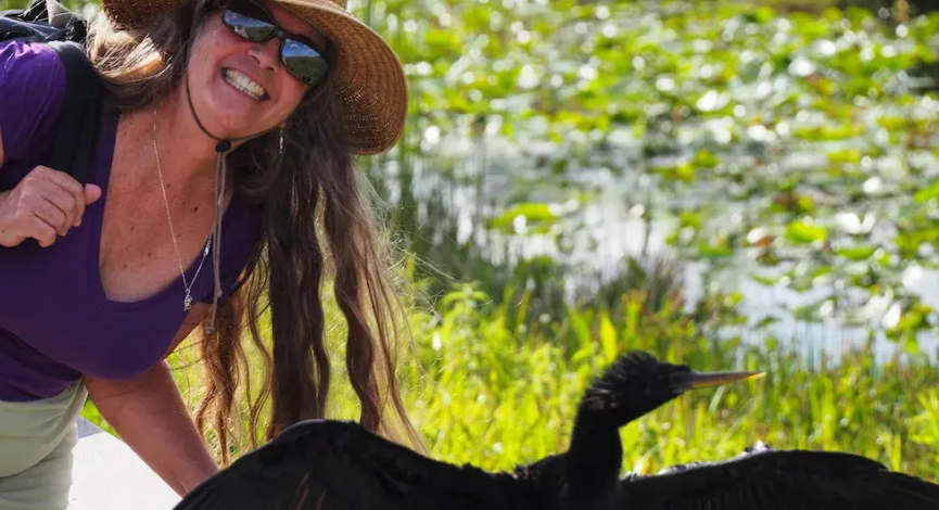 Smiling woman in sunglasses and hat next to a cormorant with outstretched wings.